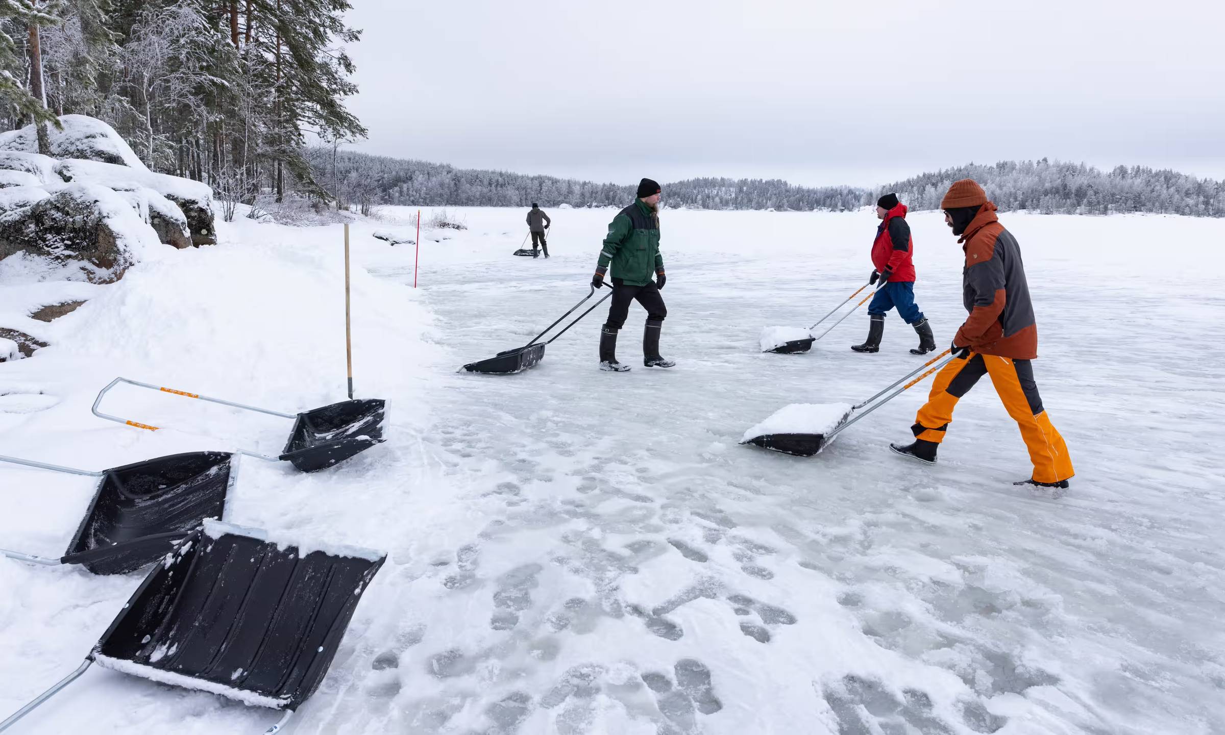 Four people dressed in outdoor winter gear shoveling snow from the middle of a body of water to the banks
