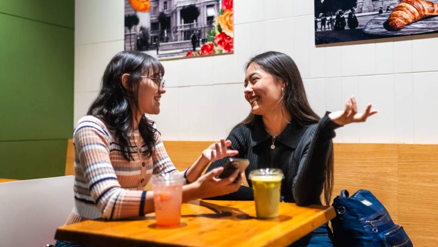 Two women sit at a wooden table facing each other, both smiling and gesturing while talking. One wears a brown and white striped cardigan with glasses; the other wears a black shirt. Orange and yellow beverages sit on the table between them. Framed photographs hang on the white and green walls behind them.