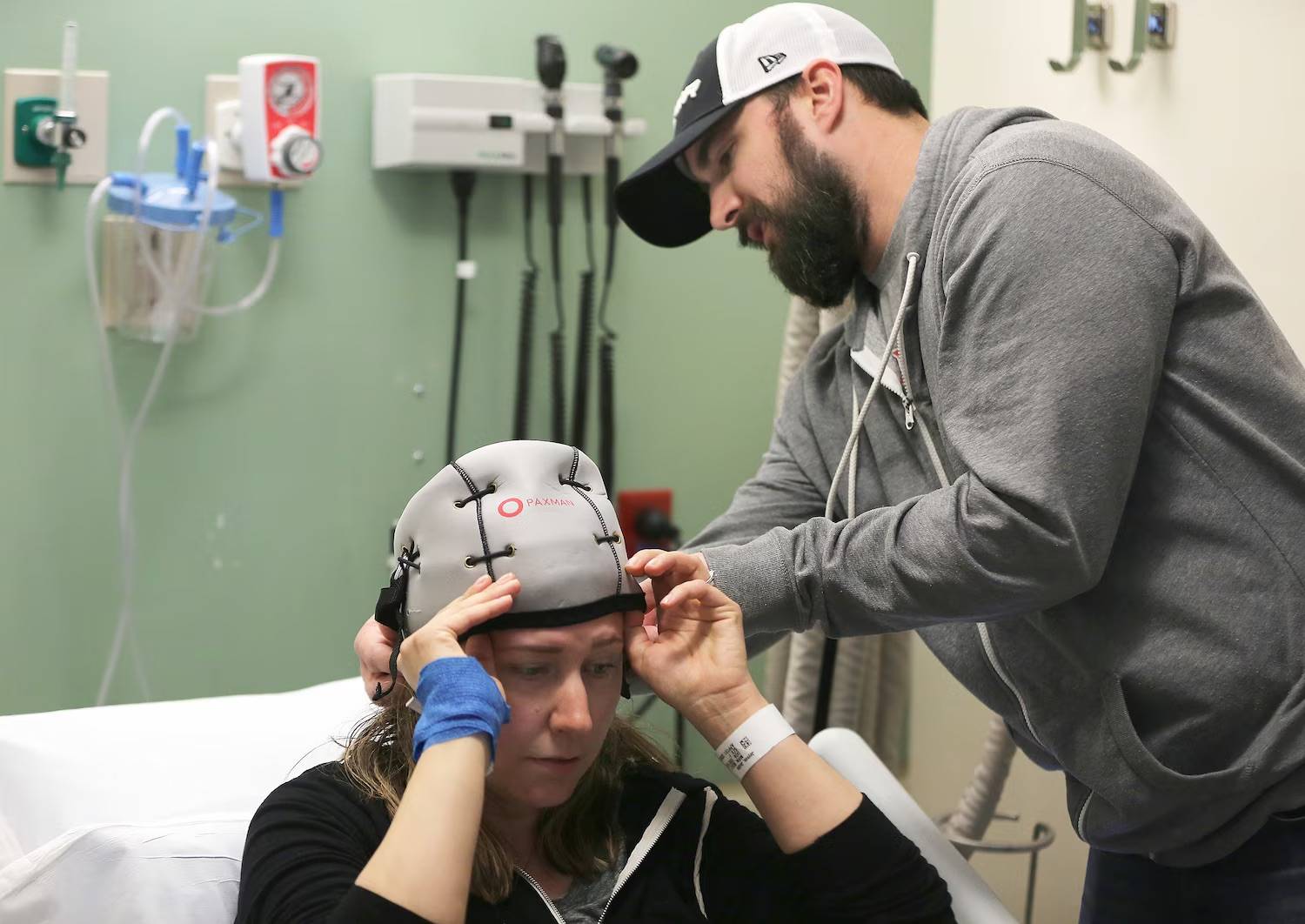Person in a grey hoodie and hat helping put a grey cooling head piece on a patient in a hospital bed