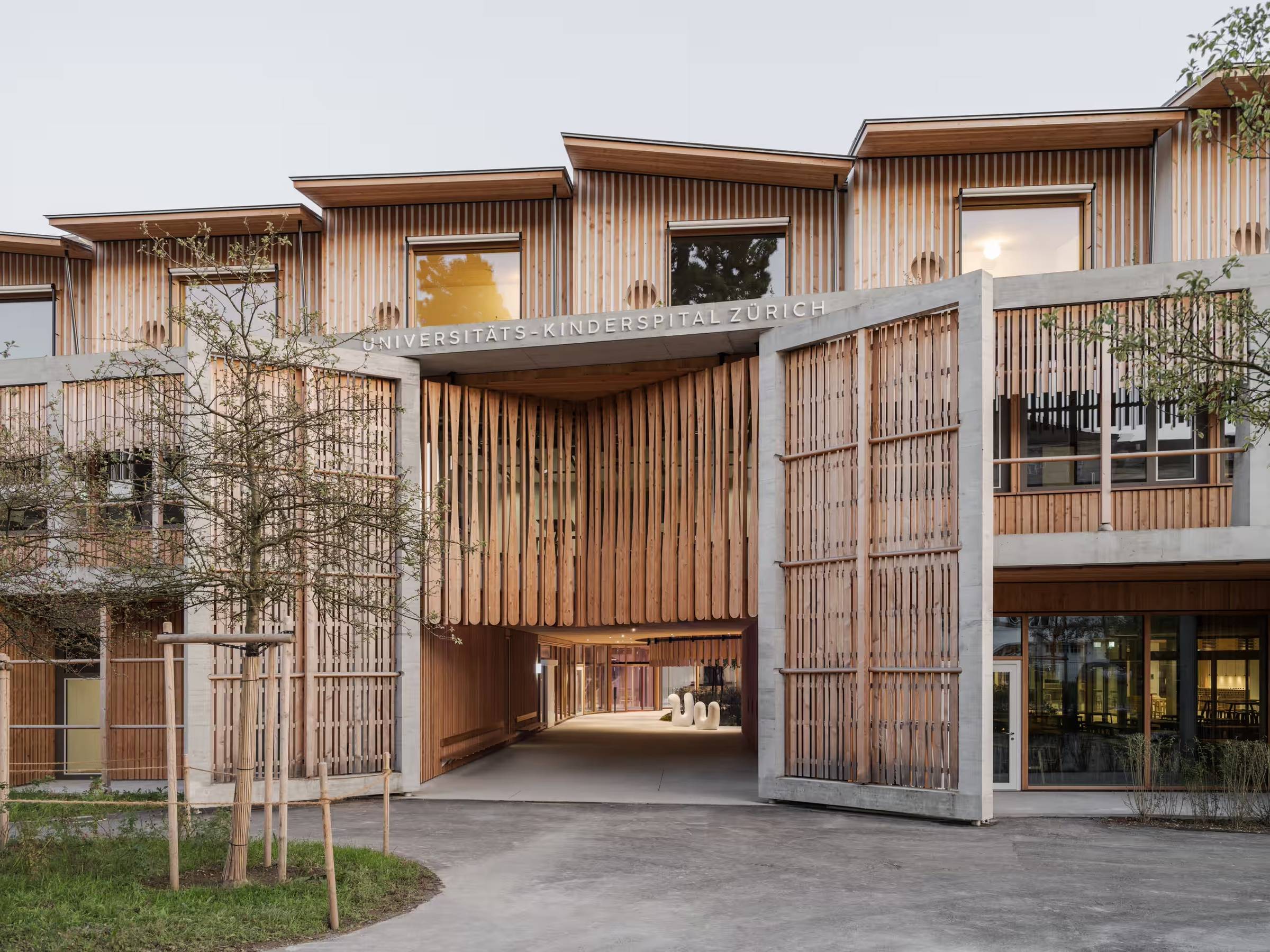 The exterior of a unique looking hospital, it has large, gate-like entry doors, wood slats on all exterior facets, windows, and a courtyard visible through the gate doors