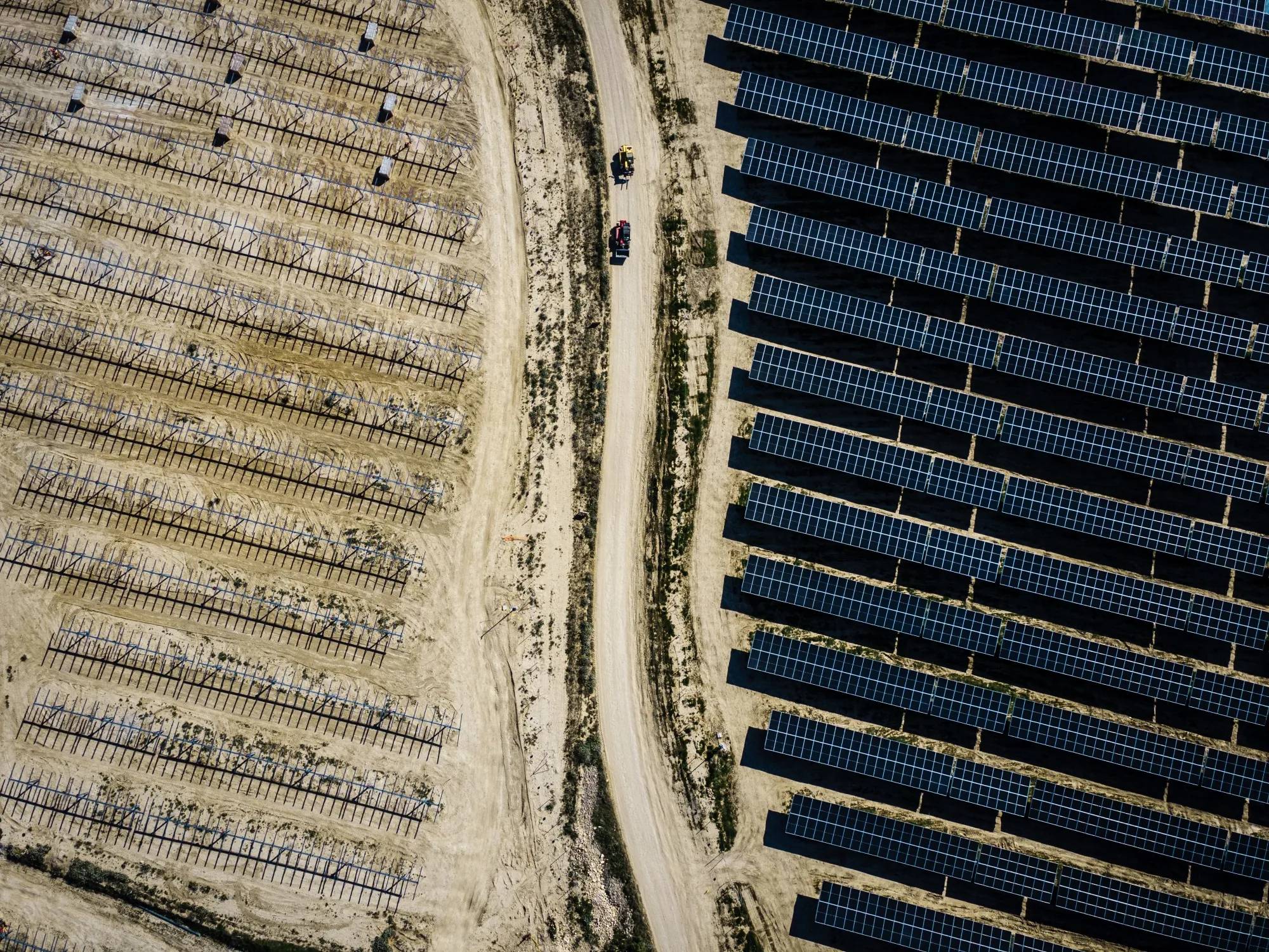 Aerial view of a solar farm on the right with dark blue panels arranged in rows, separated by a dirt road from an agricultural field on the left awaiting panels to be installed with irrigation lines and two vehicles on the road.