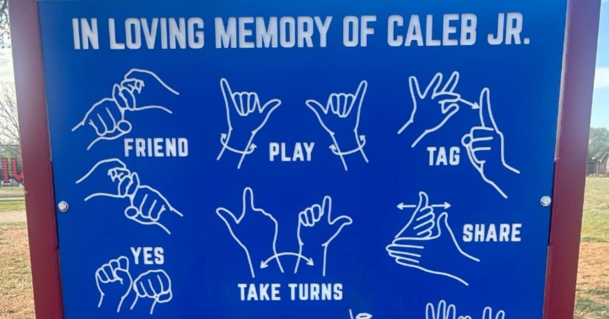A blue sign with white engravings that say "In loving memory of Caleb Jr." and depictions of hands signing words like "friend," "play" and more