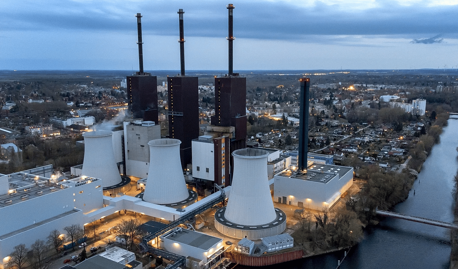 Aerial view of an industrial power plant at dusk with three tall dark chimneys, four white cooling towers, and a blue smokestack, situated beside a river with a city and forested landscape in the background.