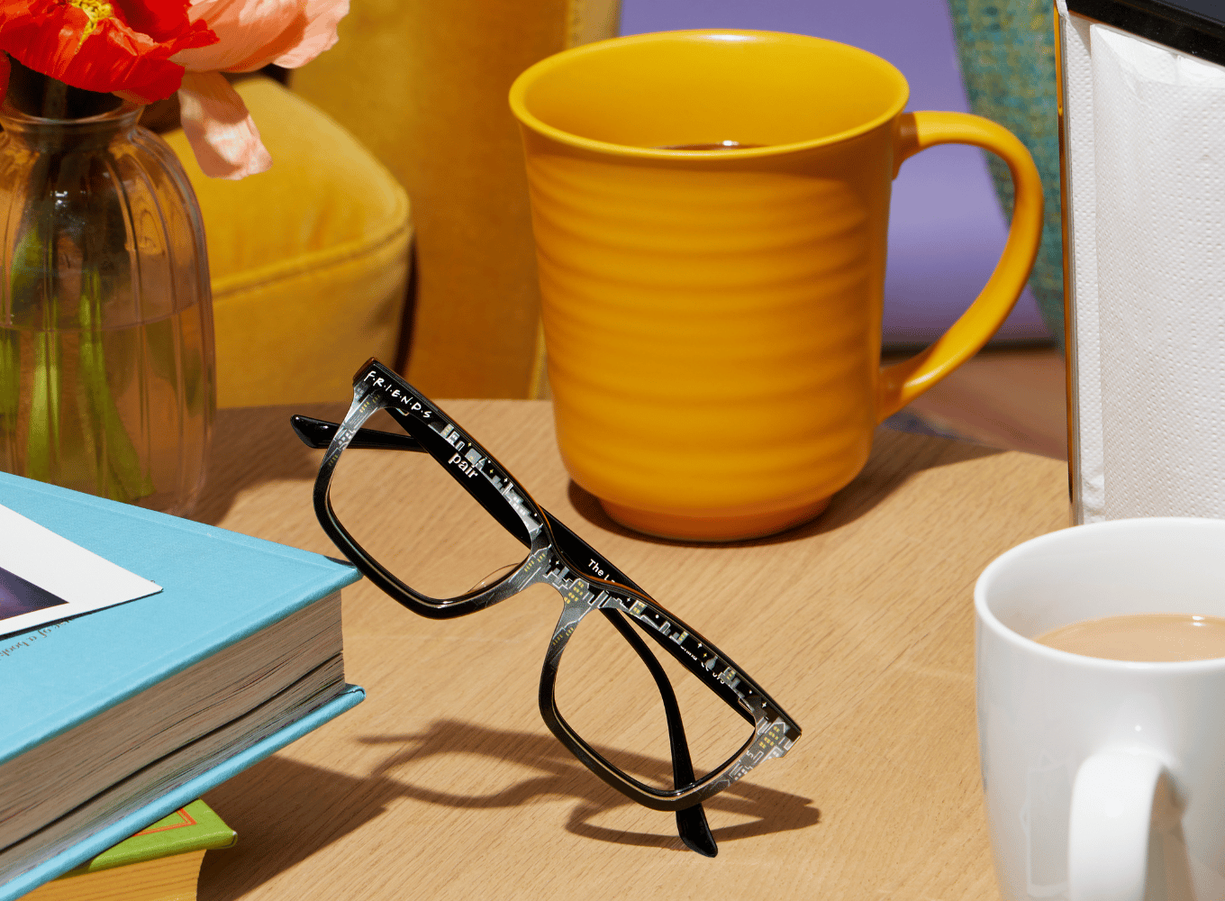 A pair of black framed glasses with a city scape in the frames resting on a blue book on a wooden table top next to two mugs of coffee