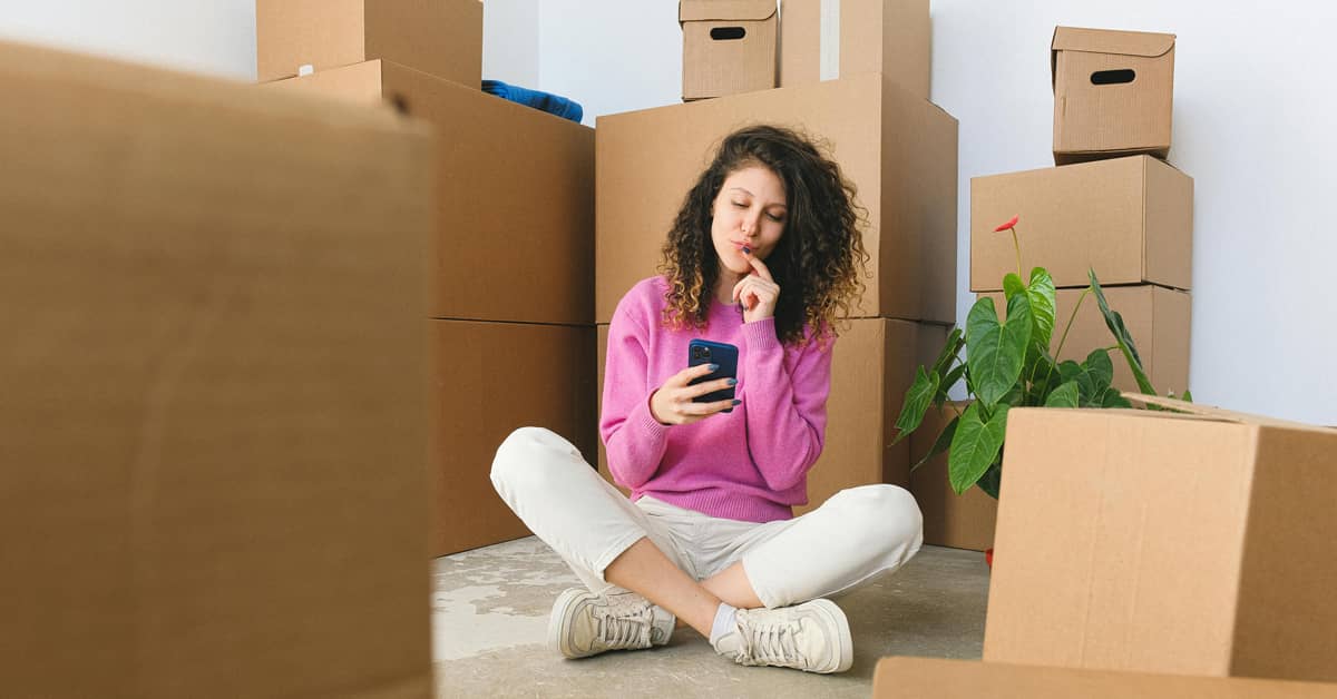 A person sitting on the floor looking at their phone surrounded by moving boxes