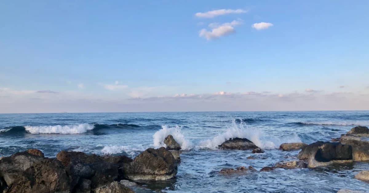 Waves crash against rocky outcrops along a coastline under a partly cloudy blue sky.
