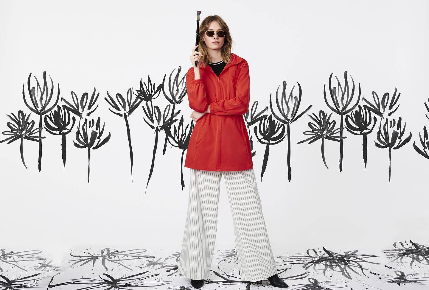 A person wearing long, pinstriped pants and a bright red rain coat, sunglasses, holding a paintbrush with black outlined flower painted on the white wall and floor behind  and under them
