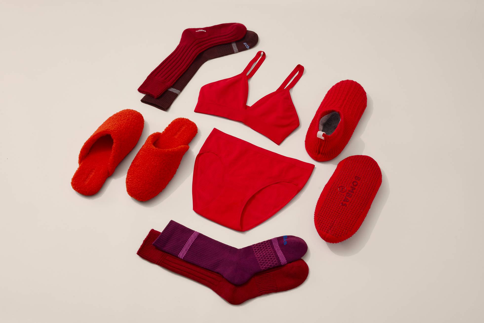 A flatlay of purple- and red-toned socks, slippers, underwear, and a bralette on a tan background