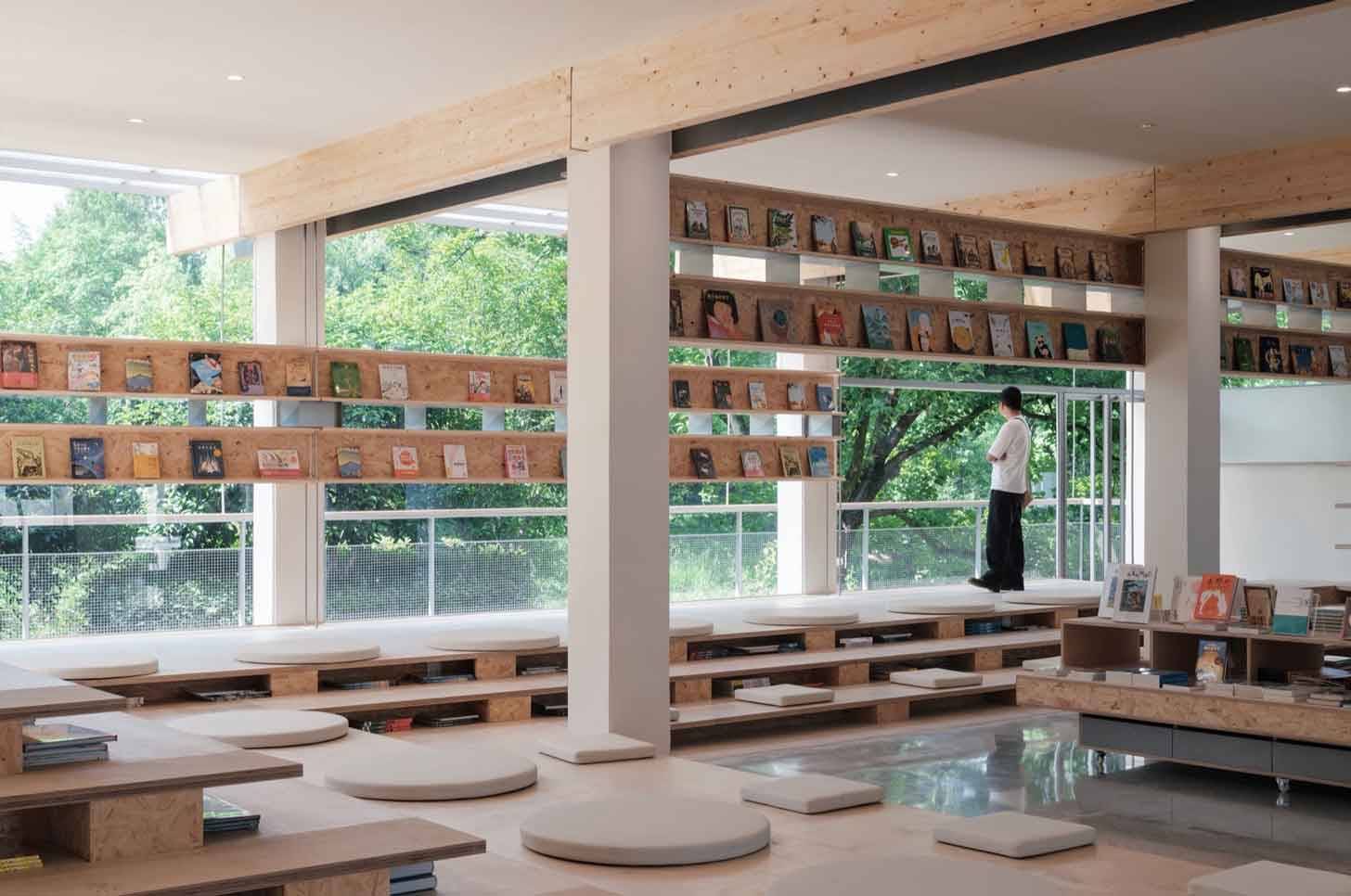 A person in front of floor-to-ceiling windows, with wooden shelves filled with books and cushions on wooden platforms in the middle of the room