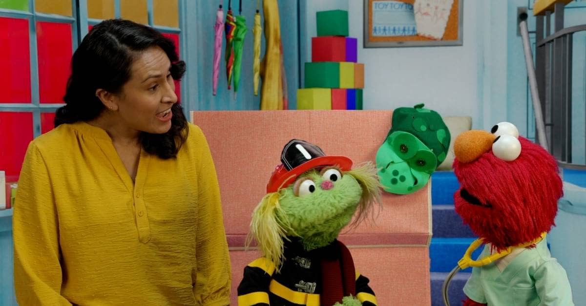 Elmo talking to a person and another muppet character on the Sesame Street set