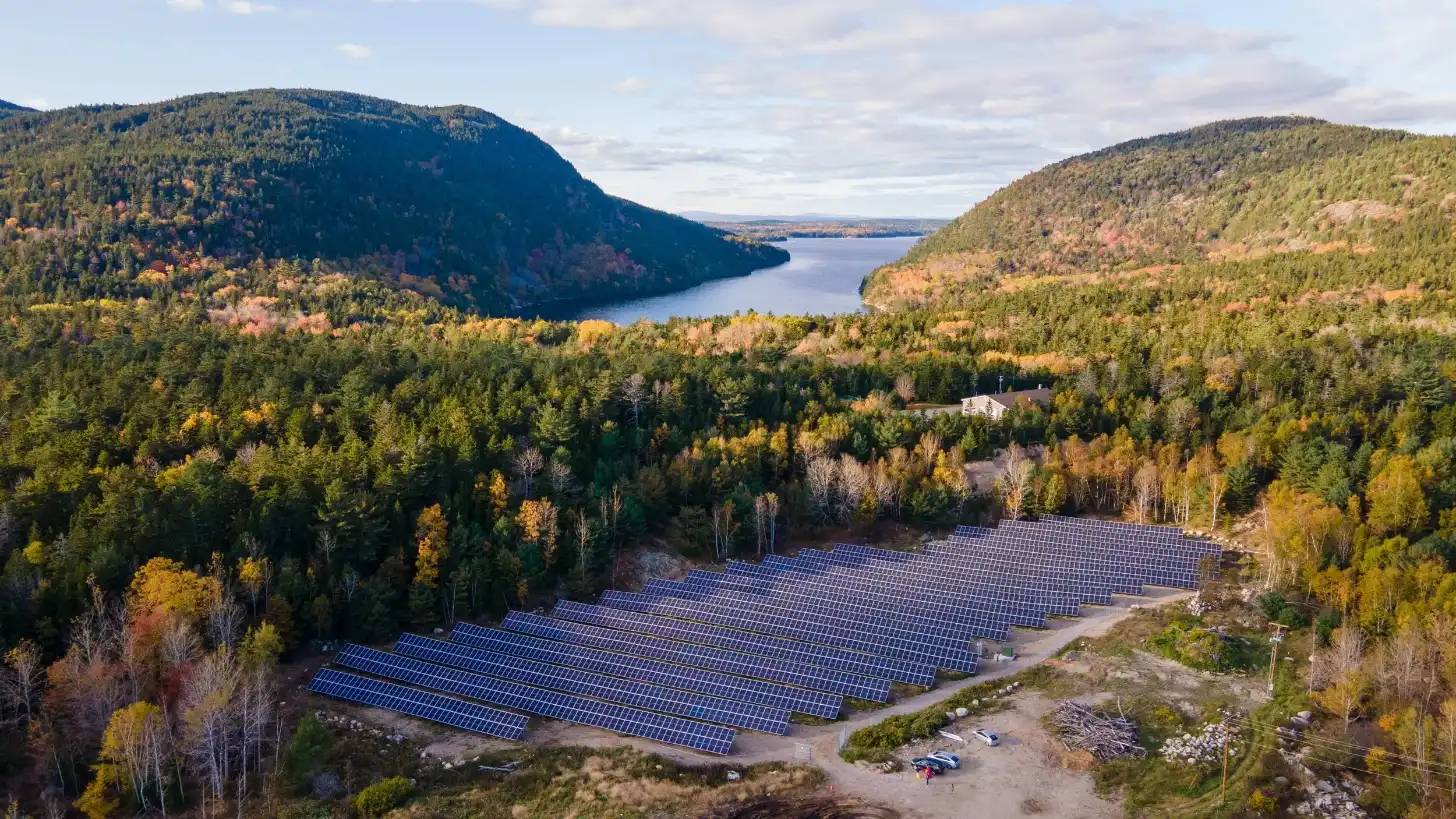 Wide-angle view of a solar array surrounded by a forest, with a body of water and tree-covered hills in the background