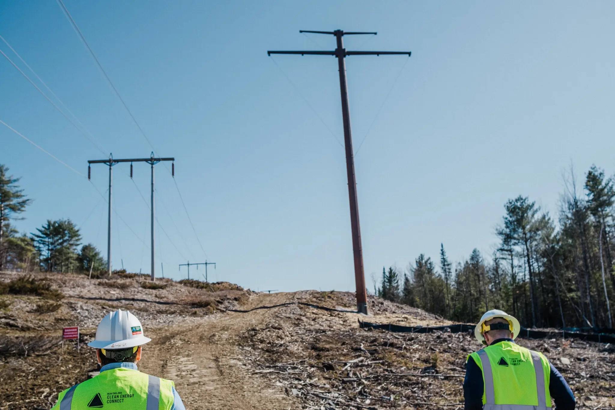 Two people in white hard hats and bright yellow vests walking up to power line poles on cleared land