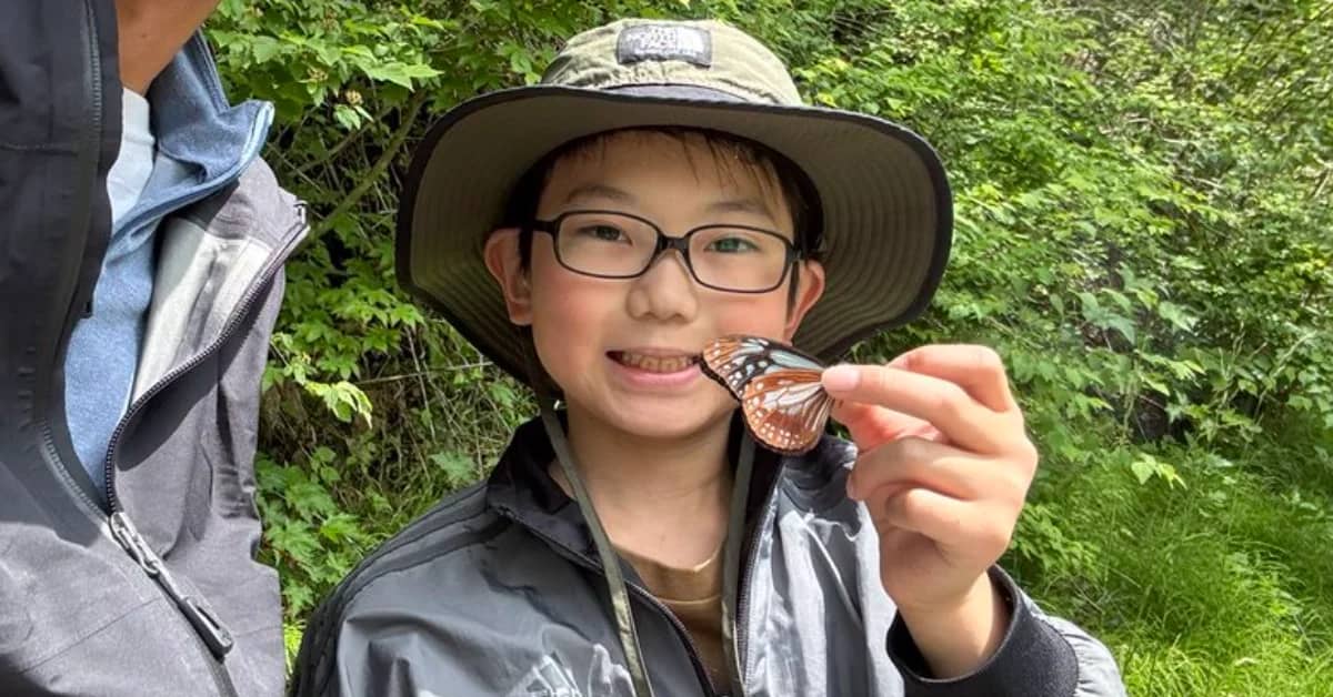 A young boy wearing a bucket hat and black rimmed glasses holding a butterfly to the camera, he's smiling