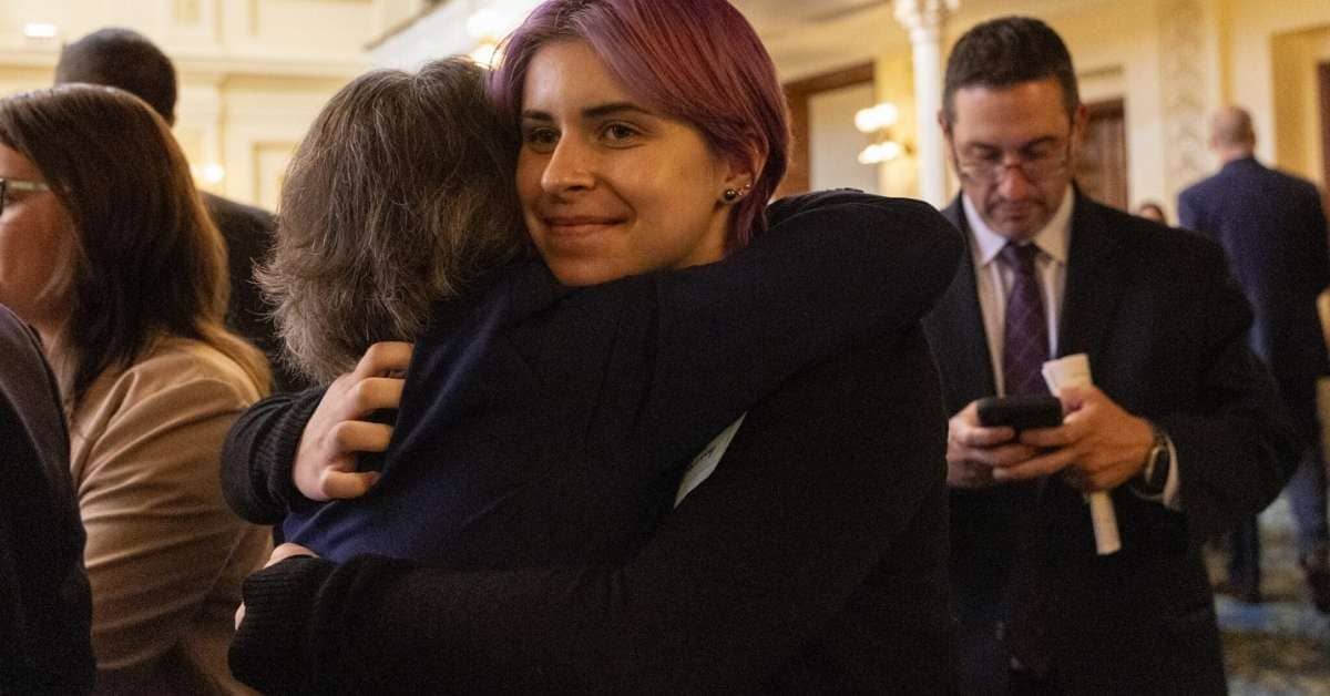 A young person hugs another in a courthouse setting as they smile. A man in the background in glasses looks at his phone.