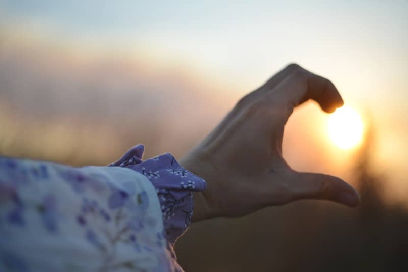 Hand forms heart shape with sun at sunset