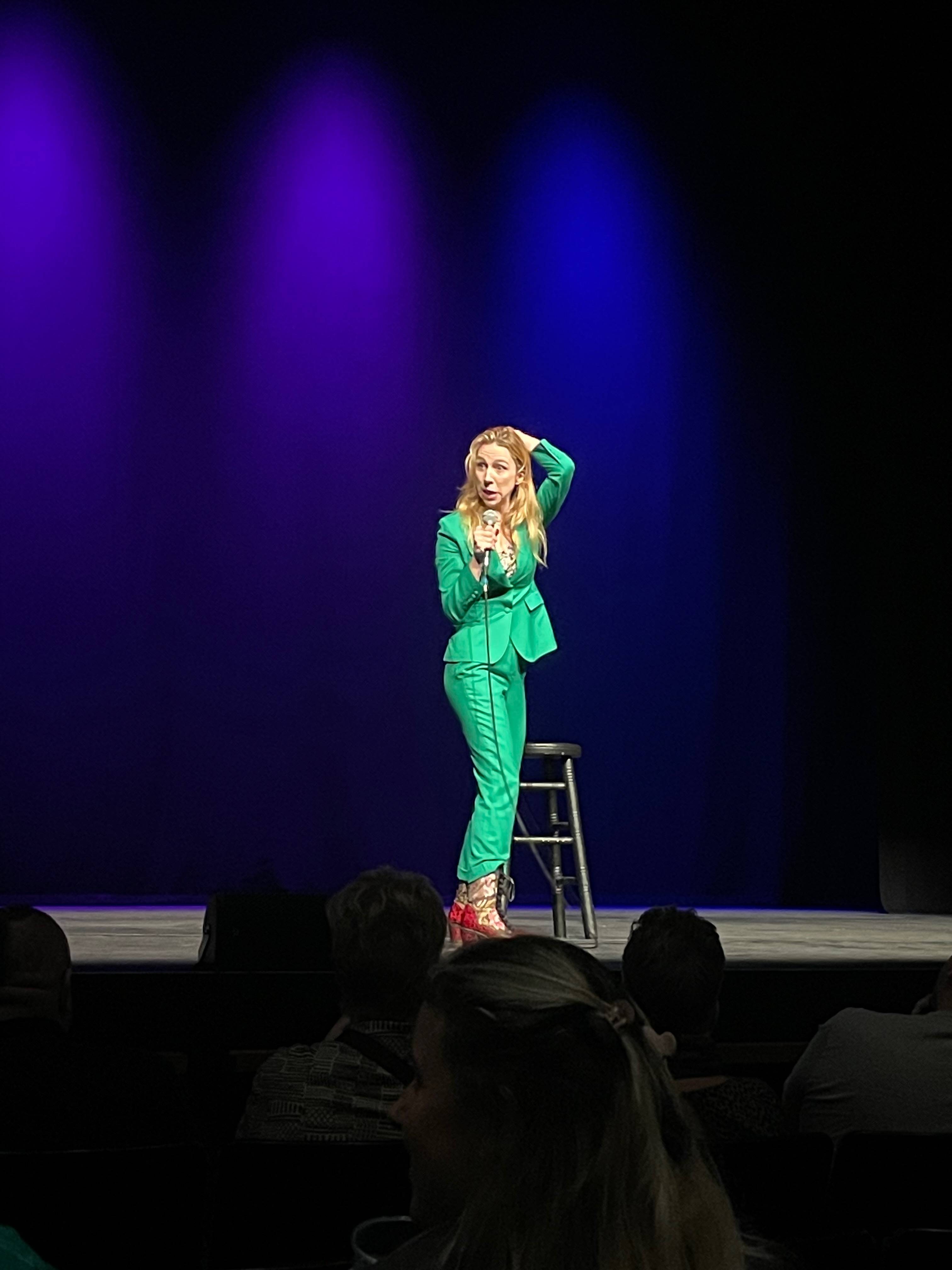 Tina Friml is onstage wearing a beautiful green suit and red and gold boots. She's a white woman with long auburn hair, holding a microphone and sweeping her hair back with one hand. She's mid-anecdote and making me laugh.