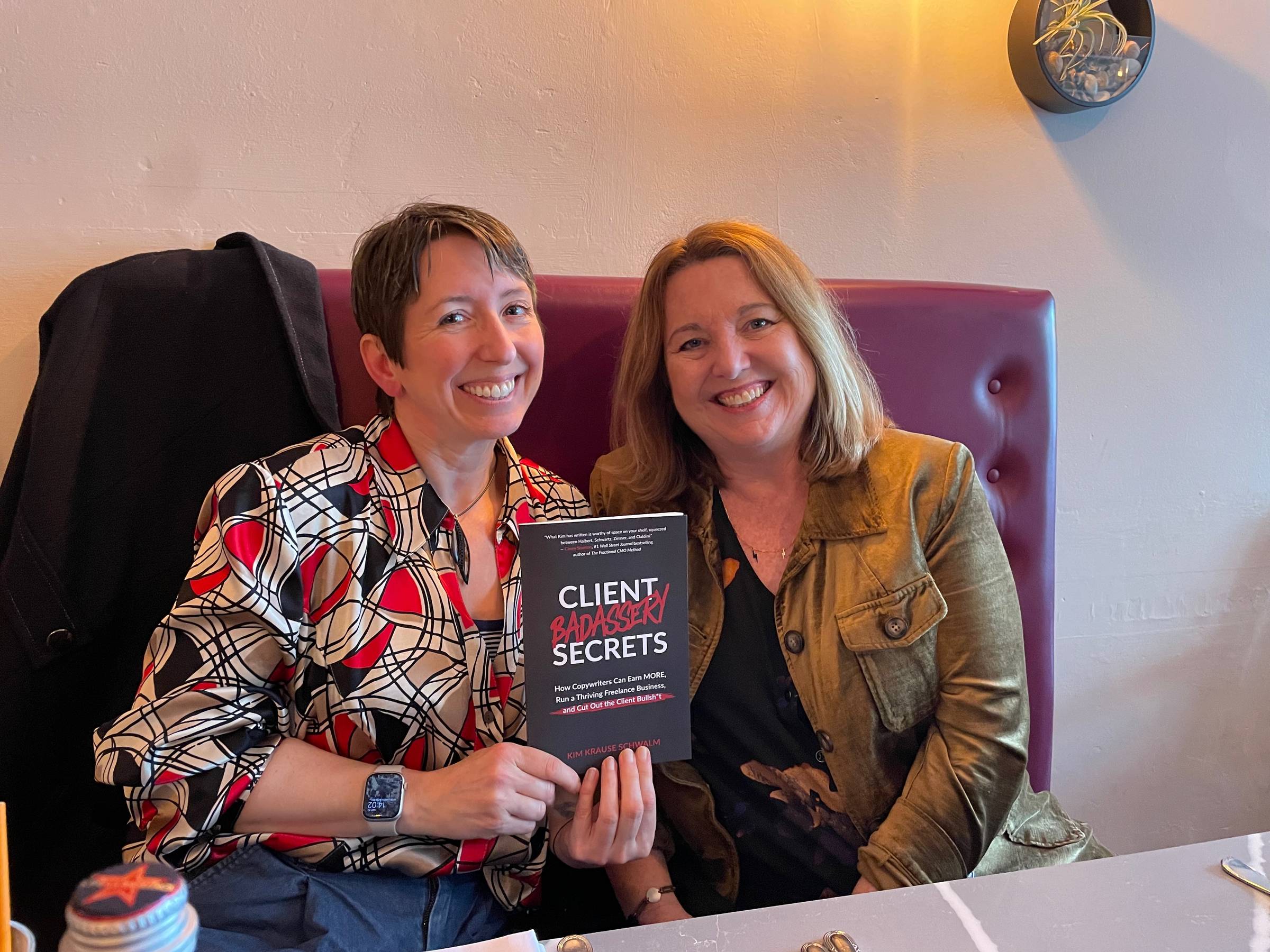Vicky and Kim are two white women in a restaurant, grinning hugely at the camera. Vicky is holding Kim’s book “Client Badassery Secrets” and looking super proud because she is.
