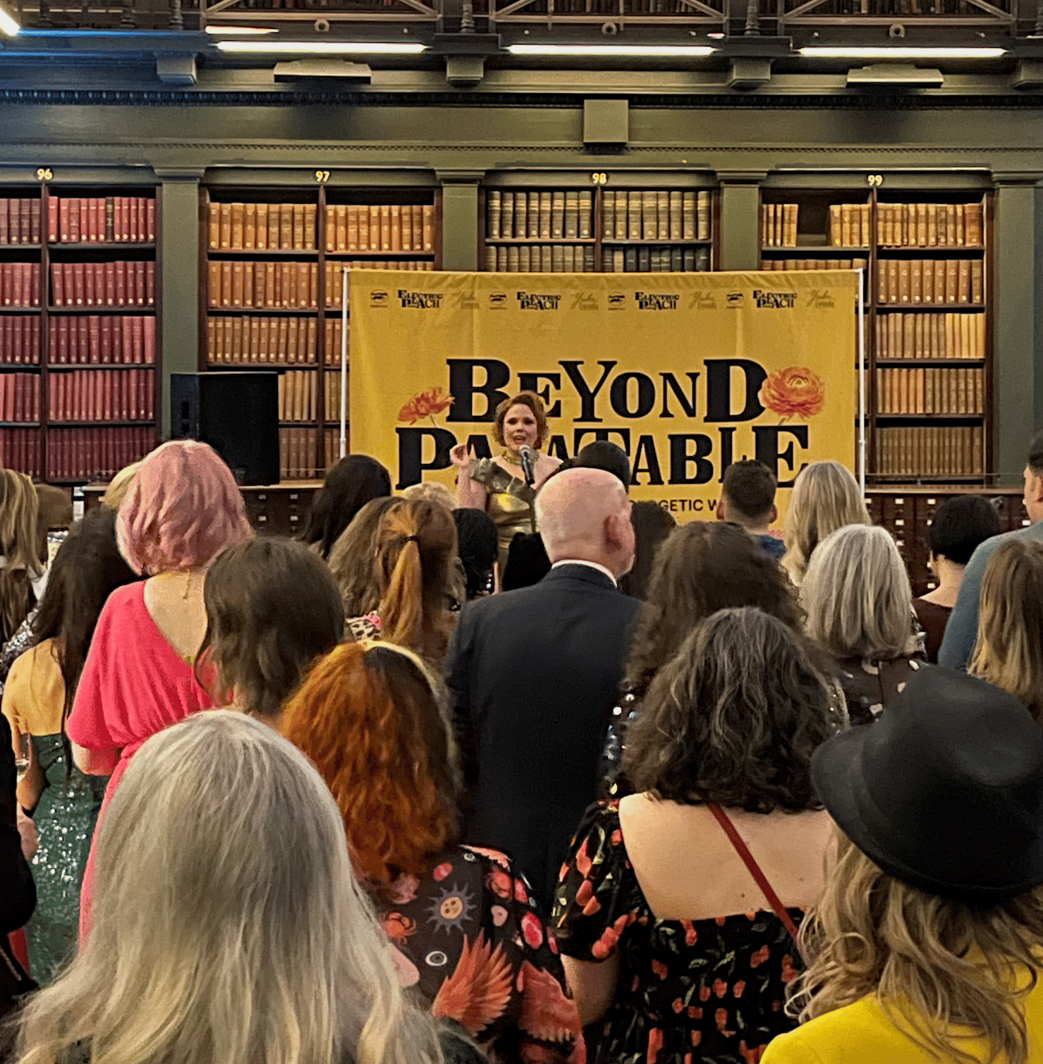 In the background, shelves of books in a fancy library painted olive green. In front of them, a huge yellow banner with “Beyond Palatable” and in front of that, a stunning redhead in a gold dress speaking into a microphone, holding up her finger. She look