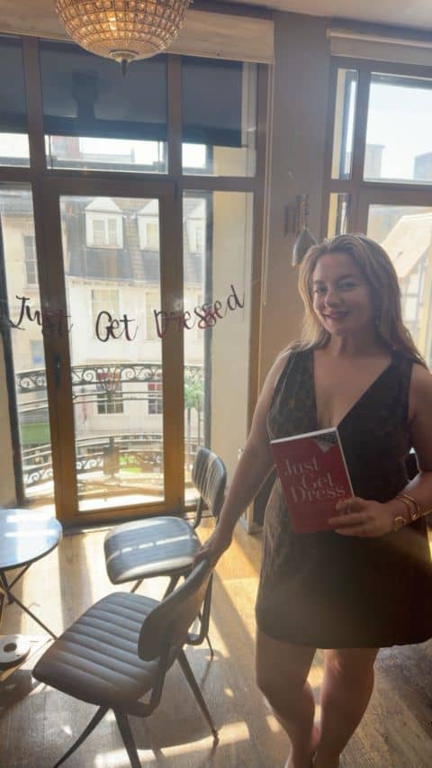 Samantha is a white woman with long dark hair, wearing a short dark dress, holding a copy of her book at her launch event. The photo is a bit dim but you can see how proud she is