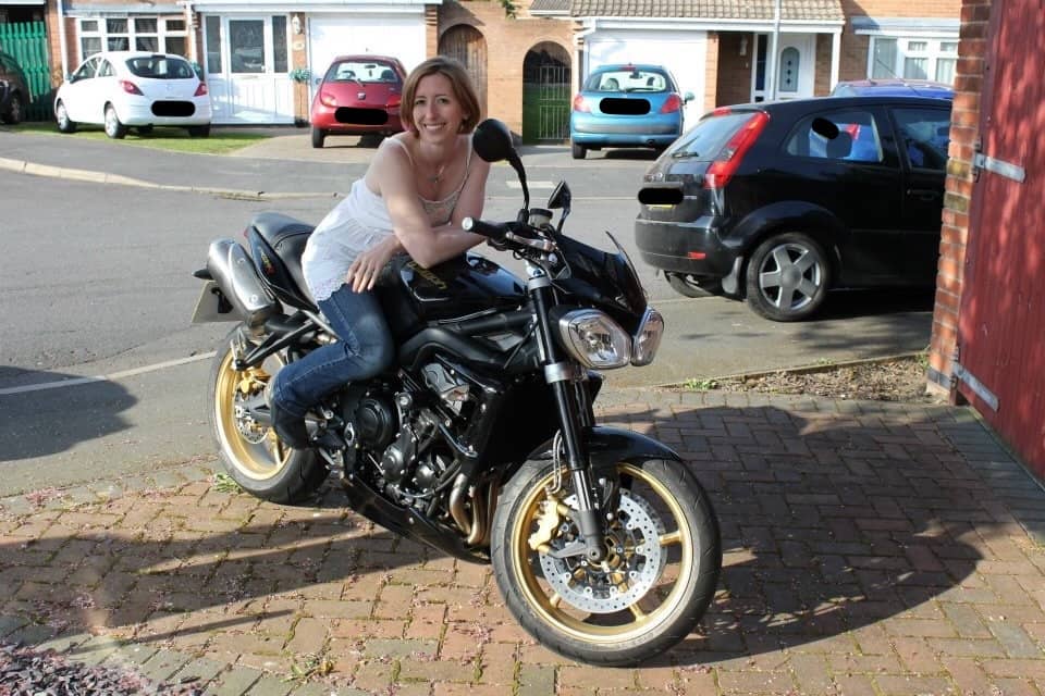 Vicky is a white woman with chin-length dark blonde hair wearing a white strappy top and jeans, sitting on a black Truimph Street Triple motorbike. She’s leaning on the tank and smiling and looking pretty darn cool and very young.