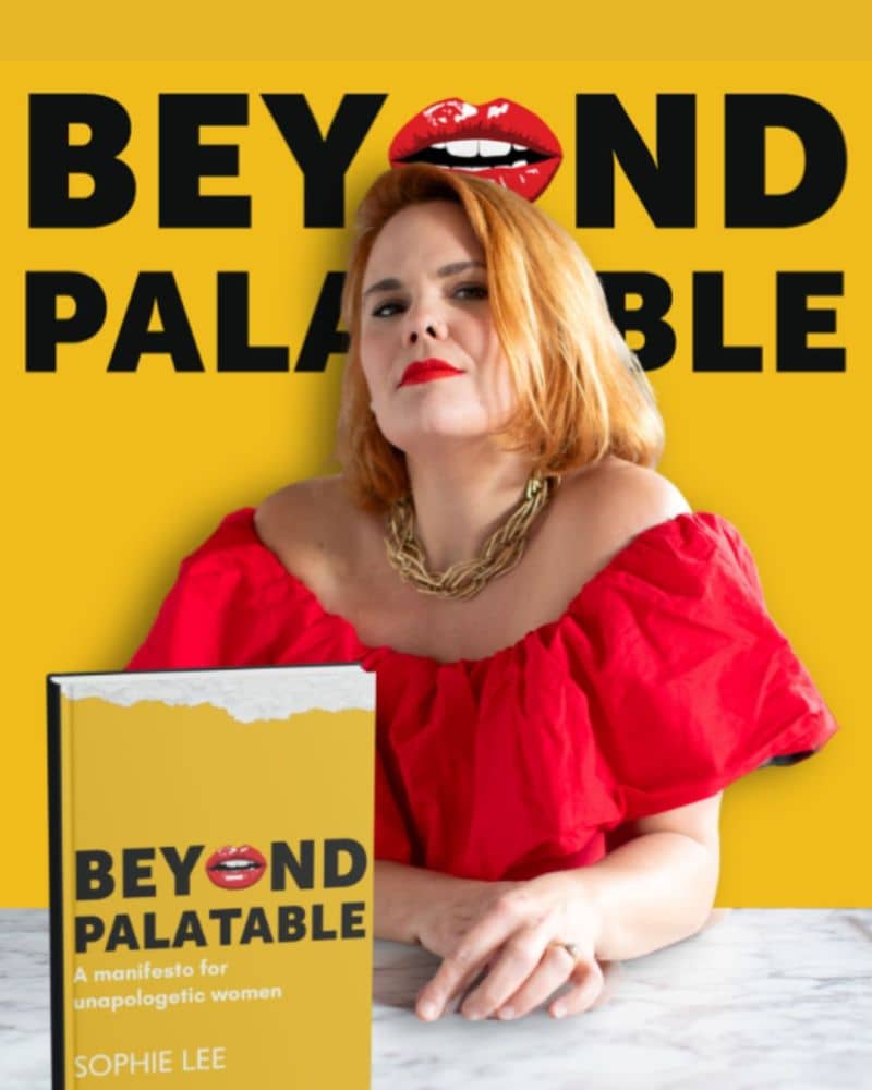 Sophie Lee has stunning chin-length red hair, red lipstick, and is staring confidently into the camera, chin up. She wears a gold rope. necklack and a red off the shoulder top. In front of her is a hardback copy of her book Beyond Palatable with its yello