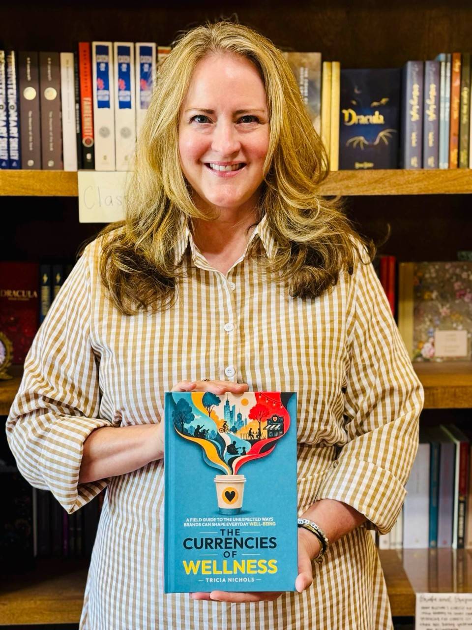 Tricia is a white woman with long dark blonde hair smiling at the camera. She’s wearing a button-down shirt with vertical stripes and holding her new book in front of her. Behind are bookshelves.