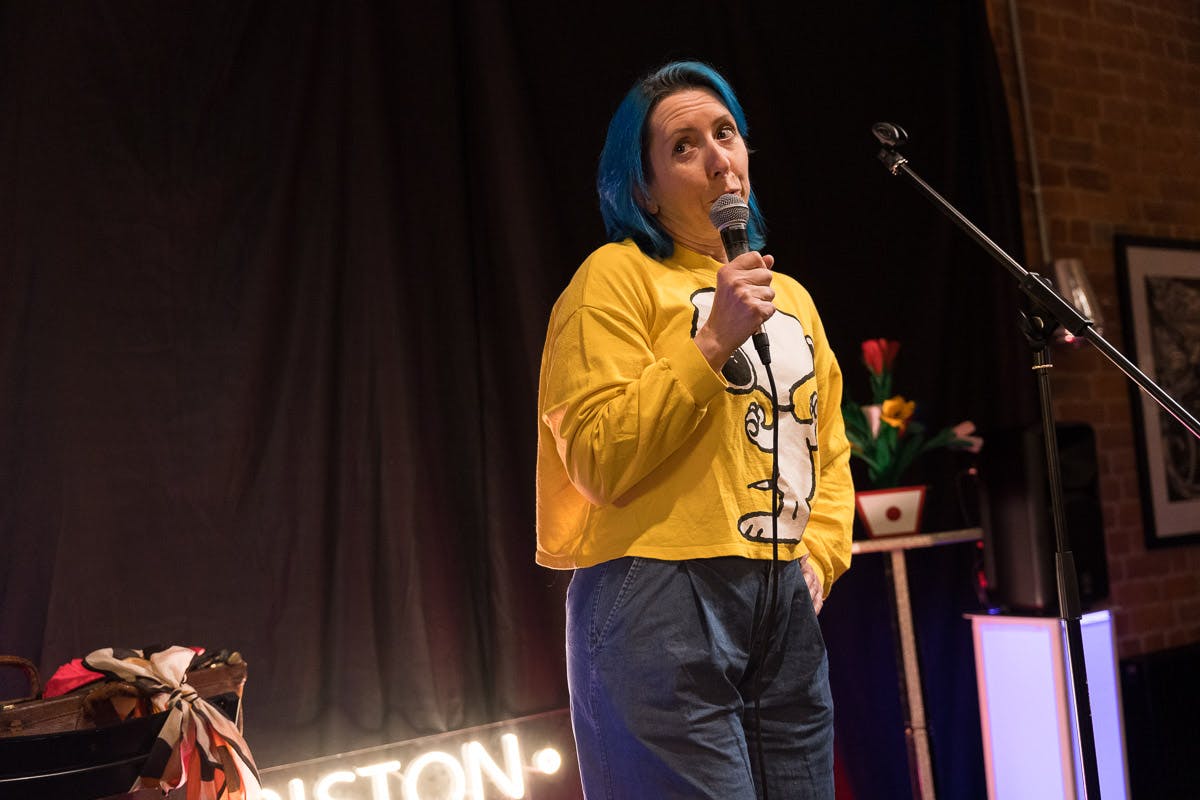 Vicky is a white woman with blue shoulder-length hair on a stage holding a microphone, looking extremely worried mid-sentence. She’s wearing blue denim trousers and a bright yellow long-sleeved t-shirt with Snoopy on it. It defnitely needs ironing but that’s because she’s been anxiously wringing it in her hands. Photo by the lovely John Cleary