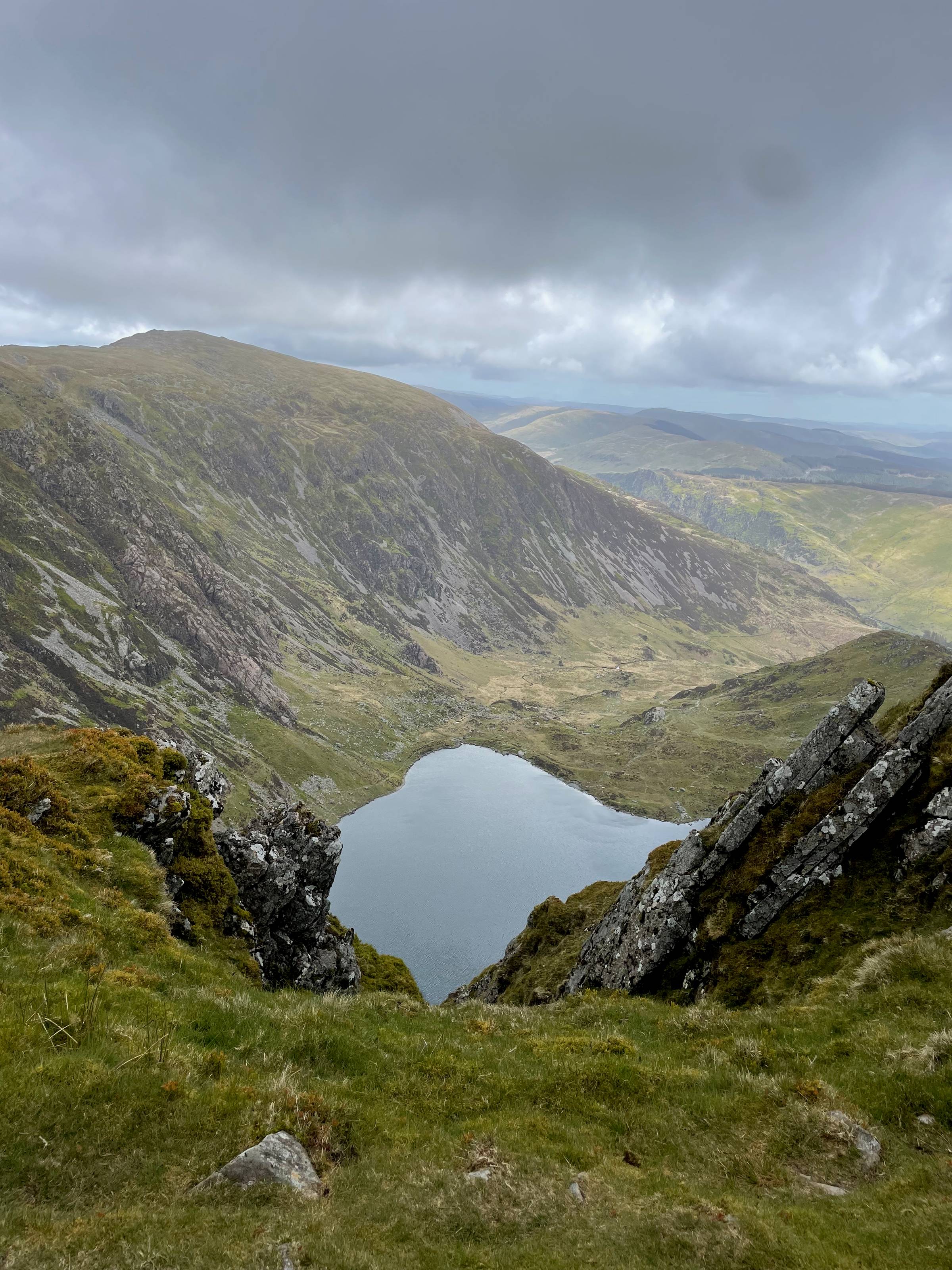 The lake from the ridge on the way to the summit of Cader Idris. Mountains and Snowdonia stretch away to the horizon. Sunlight patches the ground. Large moody clouds are overhead and we’re about to walk through one.