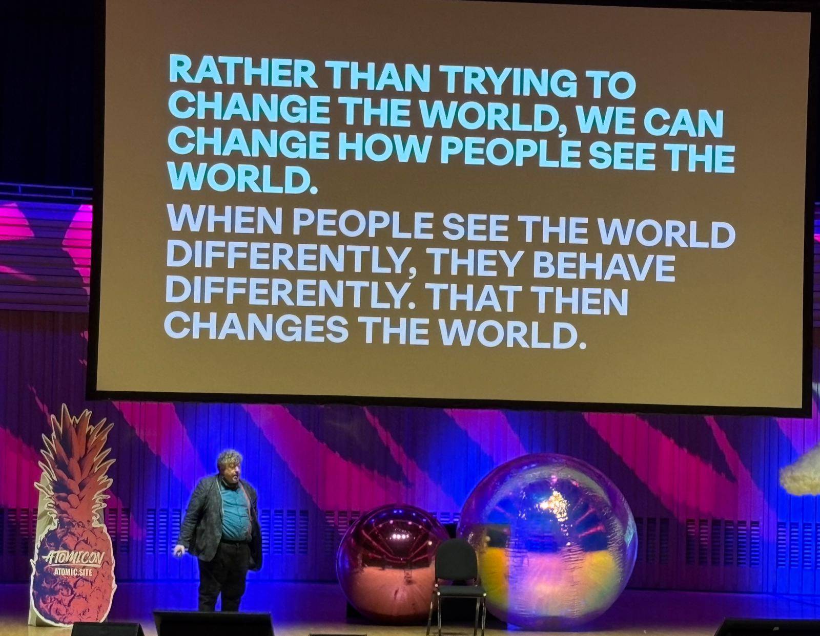 Rory Sutherland is a middle-aged white man on a stage with a cut out pineapple reading “Atomicon” and a big disco ball. Above him is a screen with a slide reading: Rather than trying to change the world, we can change how people see the world. When people