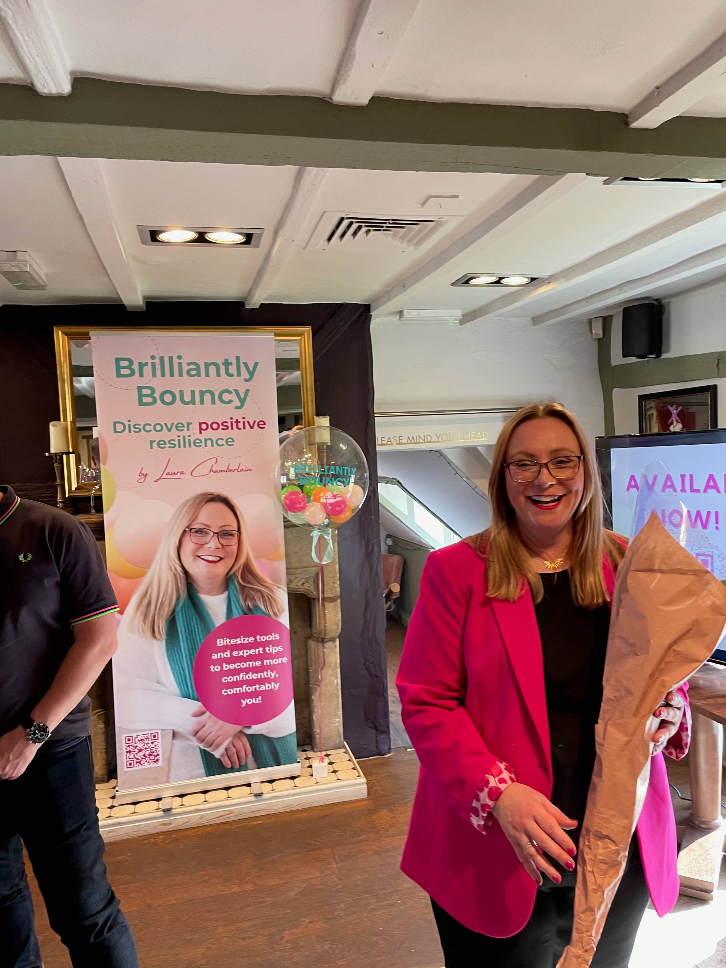 Laura is a really smiley wonderful white woman with long blonde hair and glasses. She’s wearing a stunning pink jacket with detailed cuffs and holding a bunch of flowers. Behind her is a big banner for her new book “Brilliantly Bouncy”.