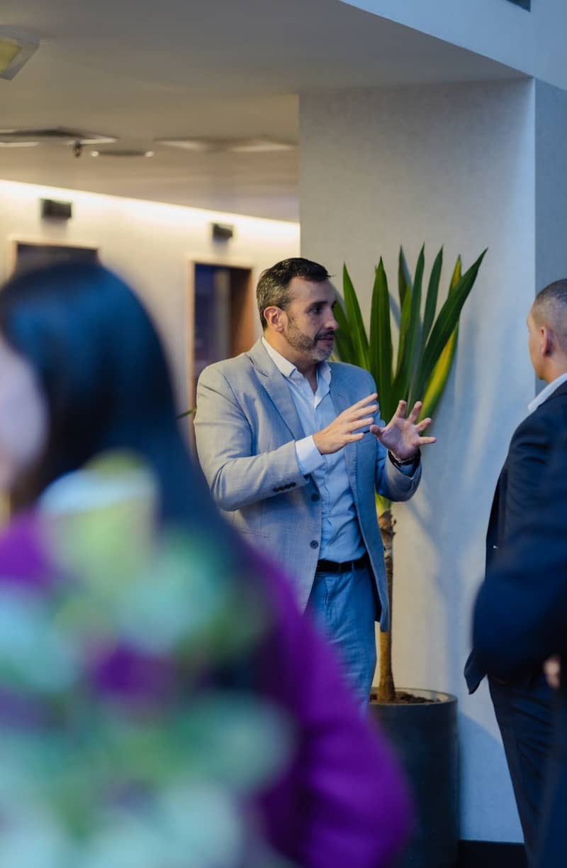 A man in a suit gestures while talking indoors.