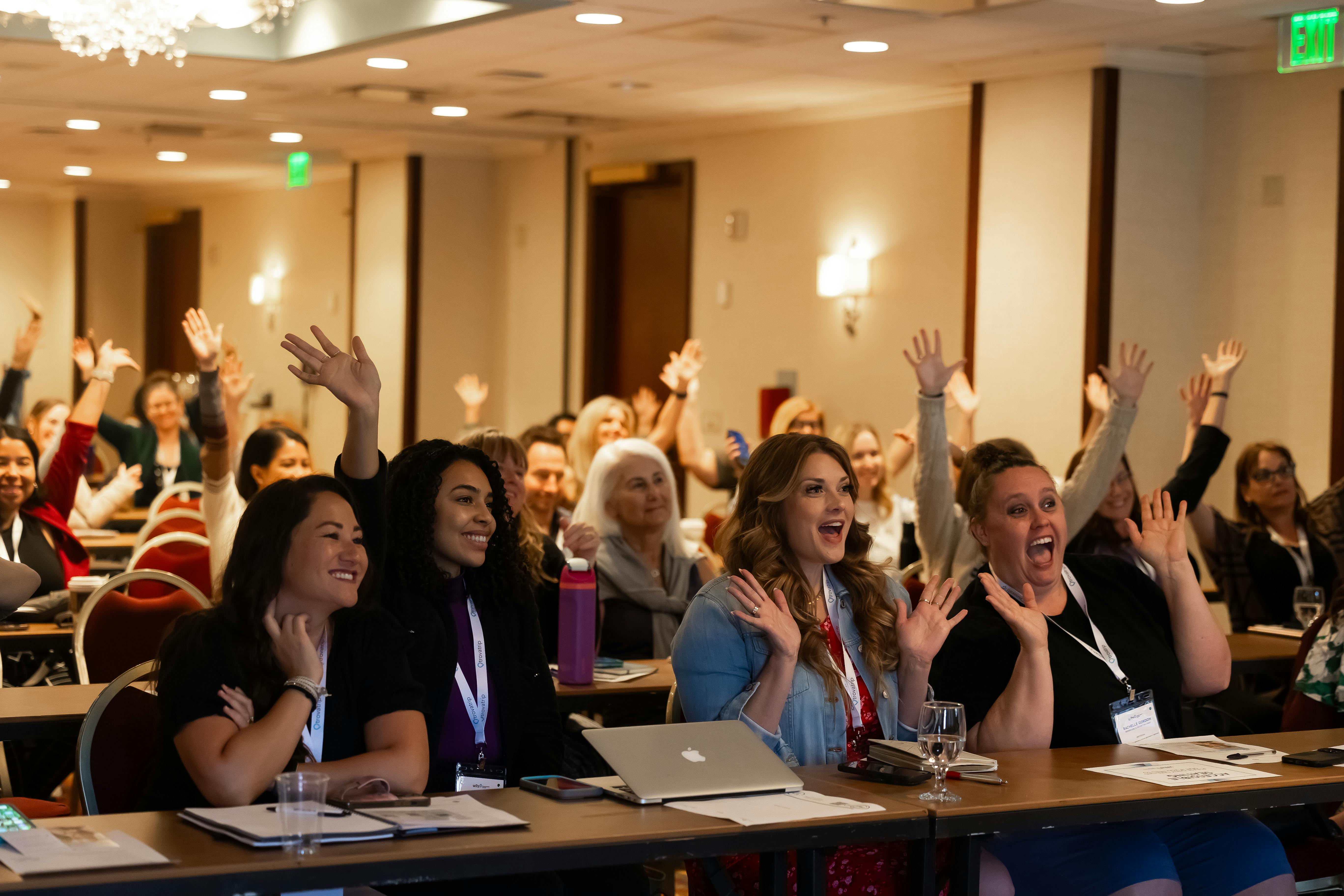 WITS Travel Creator Summit attendees hold their hands up in celebration while seated at long conference tables