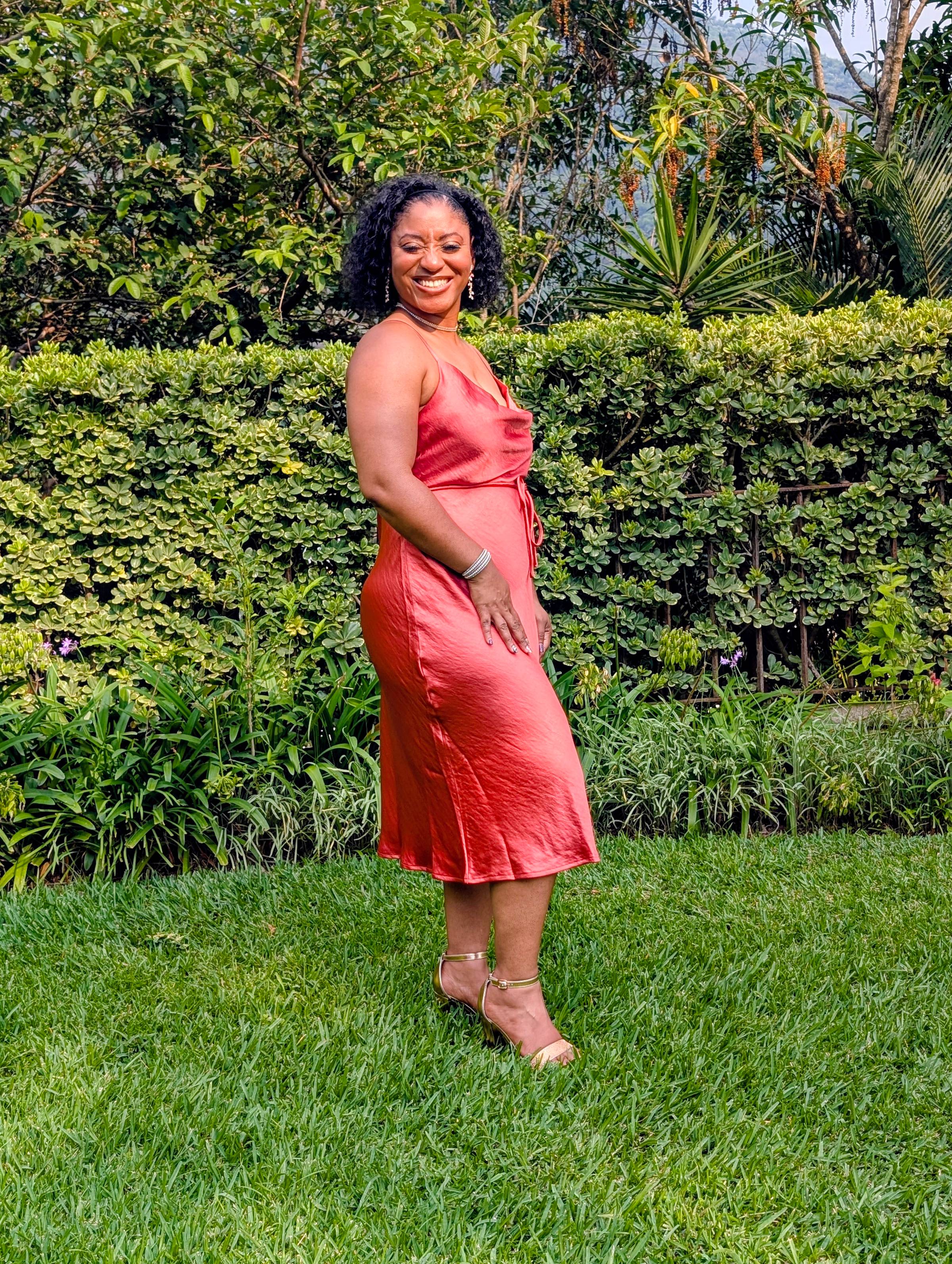 De'Christia in a rust colored strappy wedding guest dress standing in green grass with green bushes in the background.