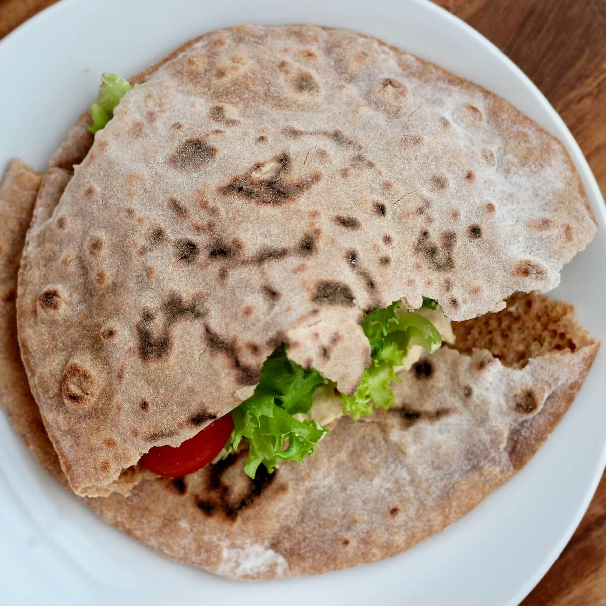 Whole wheat sourdough pita bread stuffed with lettuce, tomatoes, and hummus on a small white plate.