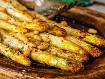Roasted white asparagus spears topped with pine nuts and cracked pepper on a rustic wooden serving dish.