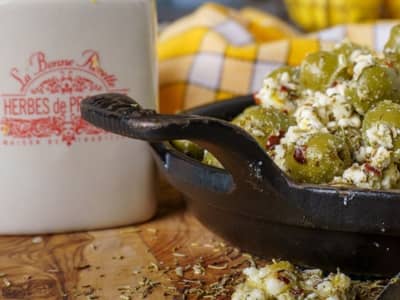 Small cast iron dish filled with green olives marinated with feta and herbs, next to a white container labeled "Herbes de Provence" on a wooden surface with a yellow plaid towel in the background.