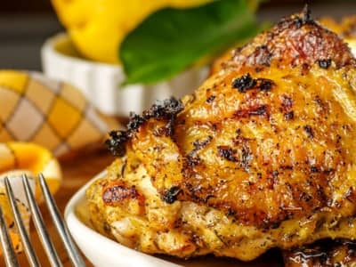 close-up of a juicy grilled chicken thigh with crispy golden skin and visible peppered seasoning, served with lemon slices and a fork on the side