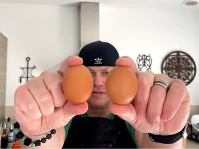 Man in a kitchen holding two brown eggs toward the camera, wearing a black hat and apron with kitchen decor in the background.