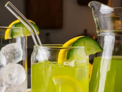 Glass of cucumber lemonade with lemon and cucumber slices, ice, and a metal straw, with a pitcher of lemonade in the background.