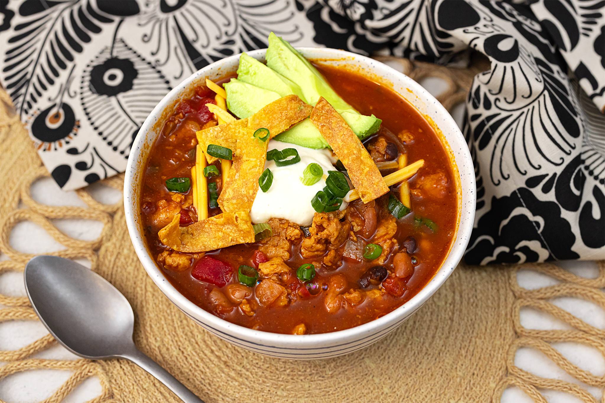 A bowl of hearty chili topped with avocado slices, sour cream, shredded cheese, and crispy tortilla strips. The dish is garnished with chopped green onions. It is placed on a woven mat with a decorative black and white cloth in the background, and a silver spoon is visible beside the bowl.