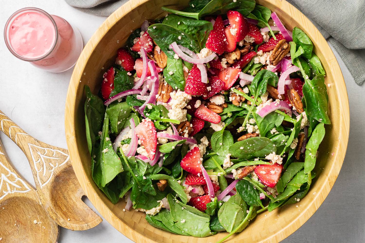 A wooden bowl filled with a fresh salad featuring spinach leaves, sliced strawberries, red onion, pecans, and crumbled cheese. To the side, there is a glass container with pink dressing and two wooden salad utensils. The setting is on a light-colored surface.