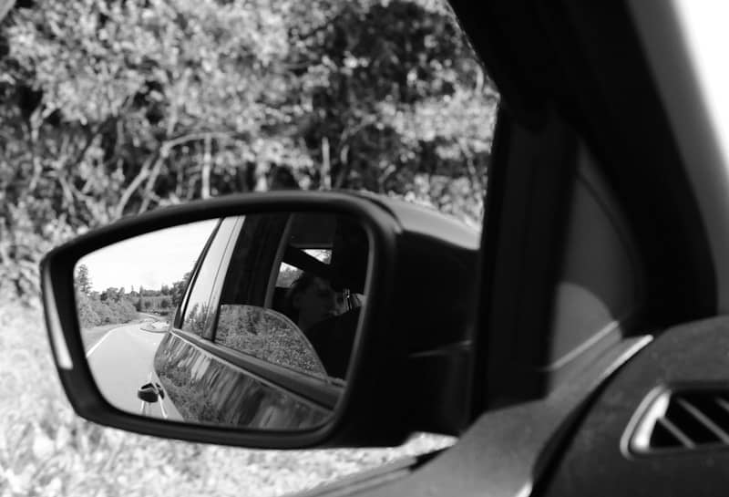 Black and white car side mirror reflecting road and trees.