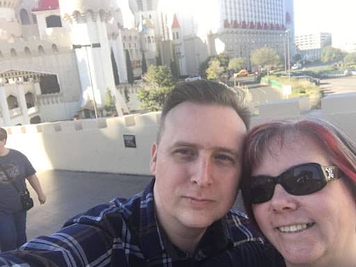 Caleb and me taking a close-up selfie outdoors in Las Vegas. Caleb, on the left, has short light brown hair and wears a blue plaid button-up shirt. I am on the right, wearing dark sunglasses, with shoulder-length dark hair featuring reddish-purple highlights. Behind us is a castle-themed hotel with white towers and red-roofed turrets, along with trees, a walkway, and nearby buildings in bright daylight.