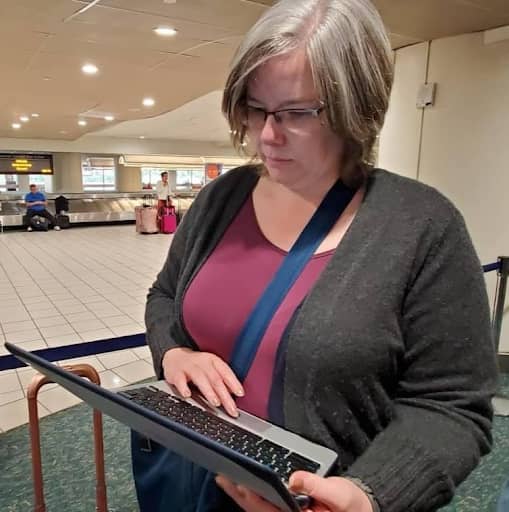 Me standing in line at an airport rental car counter, looking down while using a laptop. I have shoulder-length gray hair, wear glasses, a burgundy top, a dark gray cardigan, and a blue crossbody bag. A luggage cart handle is visible beside me, and the ba