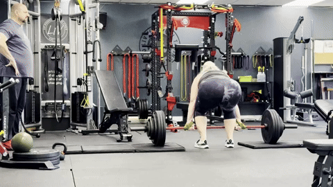 A GIF of me in athletic wear performing a deadlift in a gym. I bend down, grip the barbell loaded with large weight plates, and lift it to a standing position before lowering it back down. My trainer, Rob, stands off to the left watching (and reminding me to breathe). The gym is equipped with racks, benches, and various fitness equipment in the background.