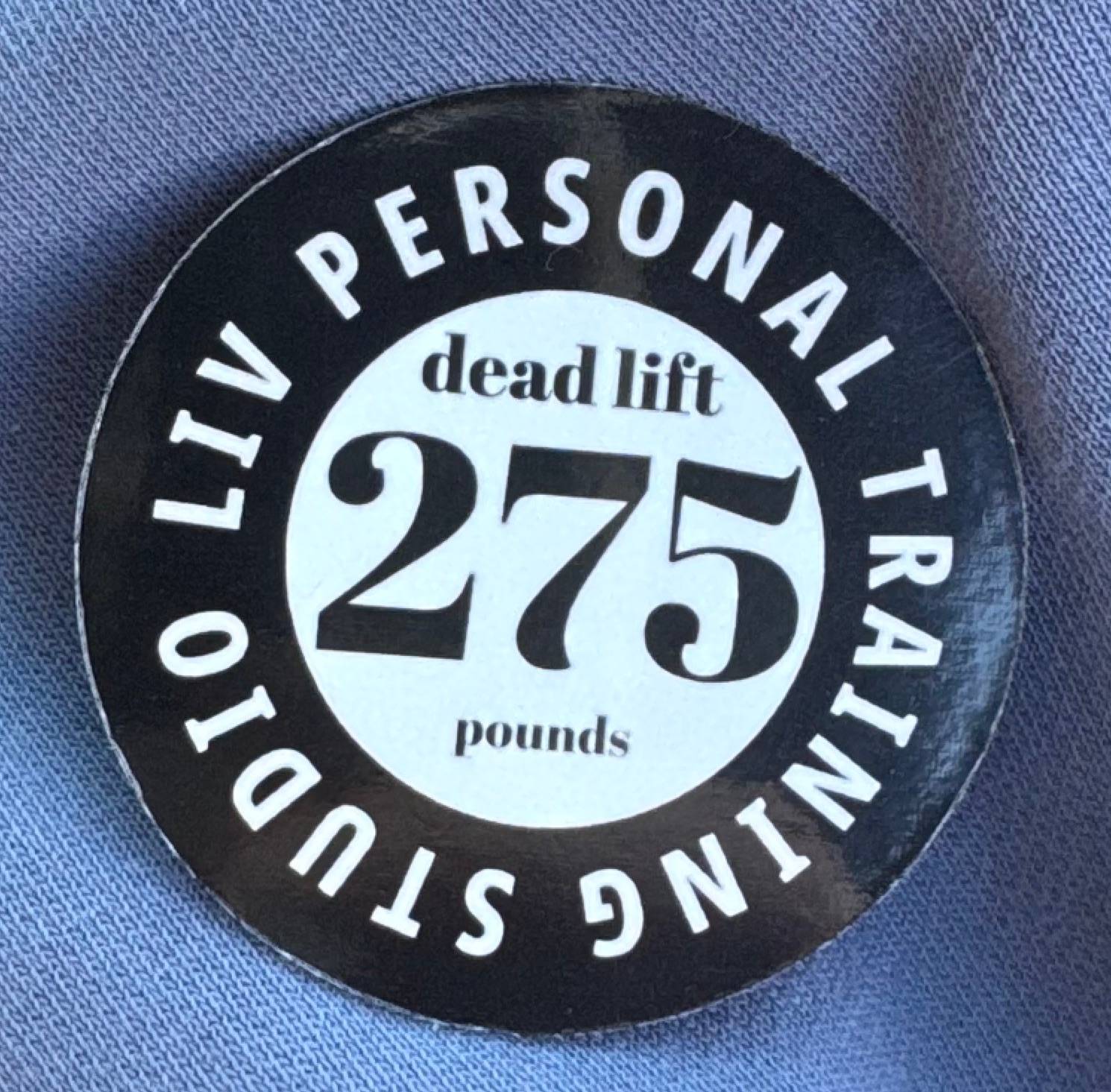 Round black and white magnet that reads 'LIV Personal Training Studio' around the outer edge. In the centre, the text says 'dead lift 275 pounds.' The magnet is placed on a light blue fabric background.