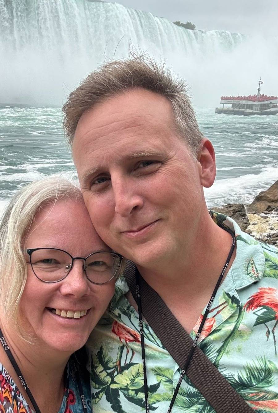 Caleb and I pose for a close-up selfie in front of Niagara Falls. He's wearing a light green tropical shirt with a flamingo and palm leaf pattern, and I'm wearing glasses and a colourful, patterned dress. In the background, the misty waterfall cascades do
