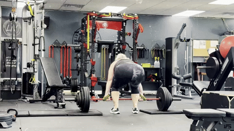 A GIF of me in athletic wear performing a deadlift in a gym. I bend down, grip the barbell loaded with large weight plates, and struggle to lift it. I barely get it off the floor before quickly putting it back down. The gym is equipped with racks, benches, and various fitness equipment in the background.