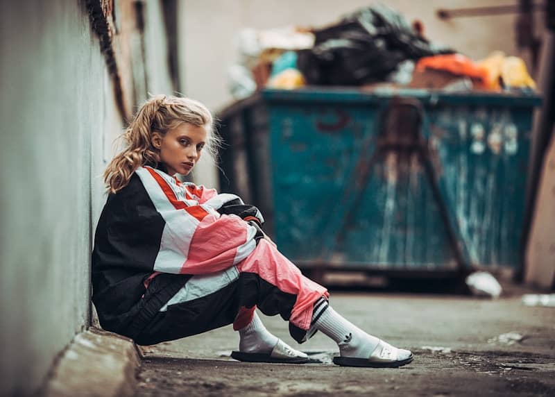 Young woman sits by wall near dumpster