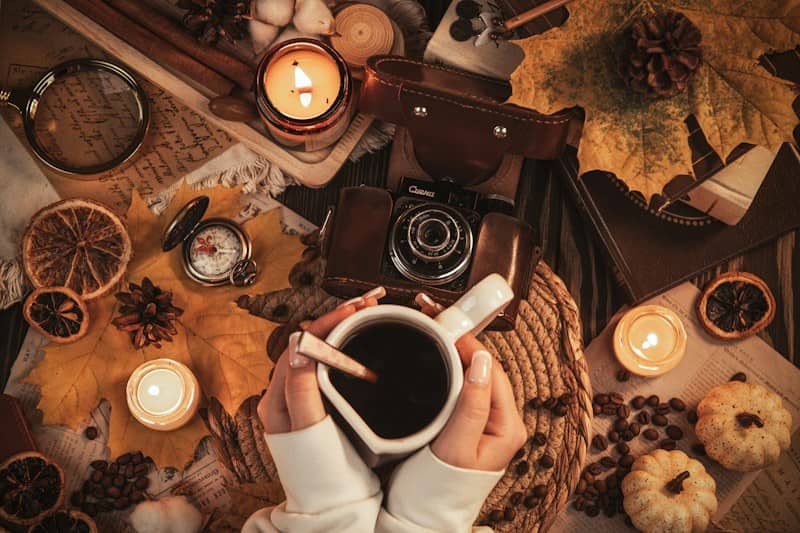 A person holding a mug of coffee surrounded by autumn leaves and candles